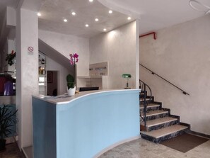 A reception area with a curved blue counter, a staircase with marble steps, and a wall-mounted control panel. - Hotel Le Ali (Rimini)