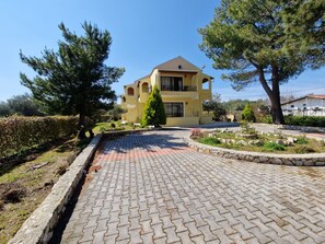 A yellow building with a paved driveway, surrounded by trees and a landscaped garden.