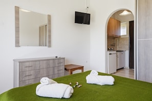 Una habitación con una cama, un aparador de madera, un televisor y una cocina visible a través de un arco.