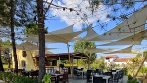Een terras buiten met tafels en stoelen, beschaduwd door grote witte luifels, omringd door plantenbakken en bomen.