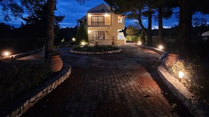 A two-story house with a paved driveway, surrounded by trees, and illuminated by exterior lights.