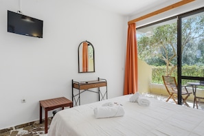A hotel room with a bed, a desk, a mirror, a TV, and a view of greenery.