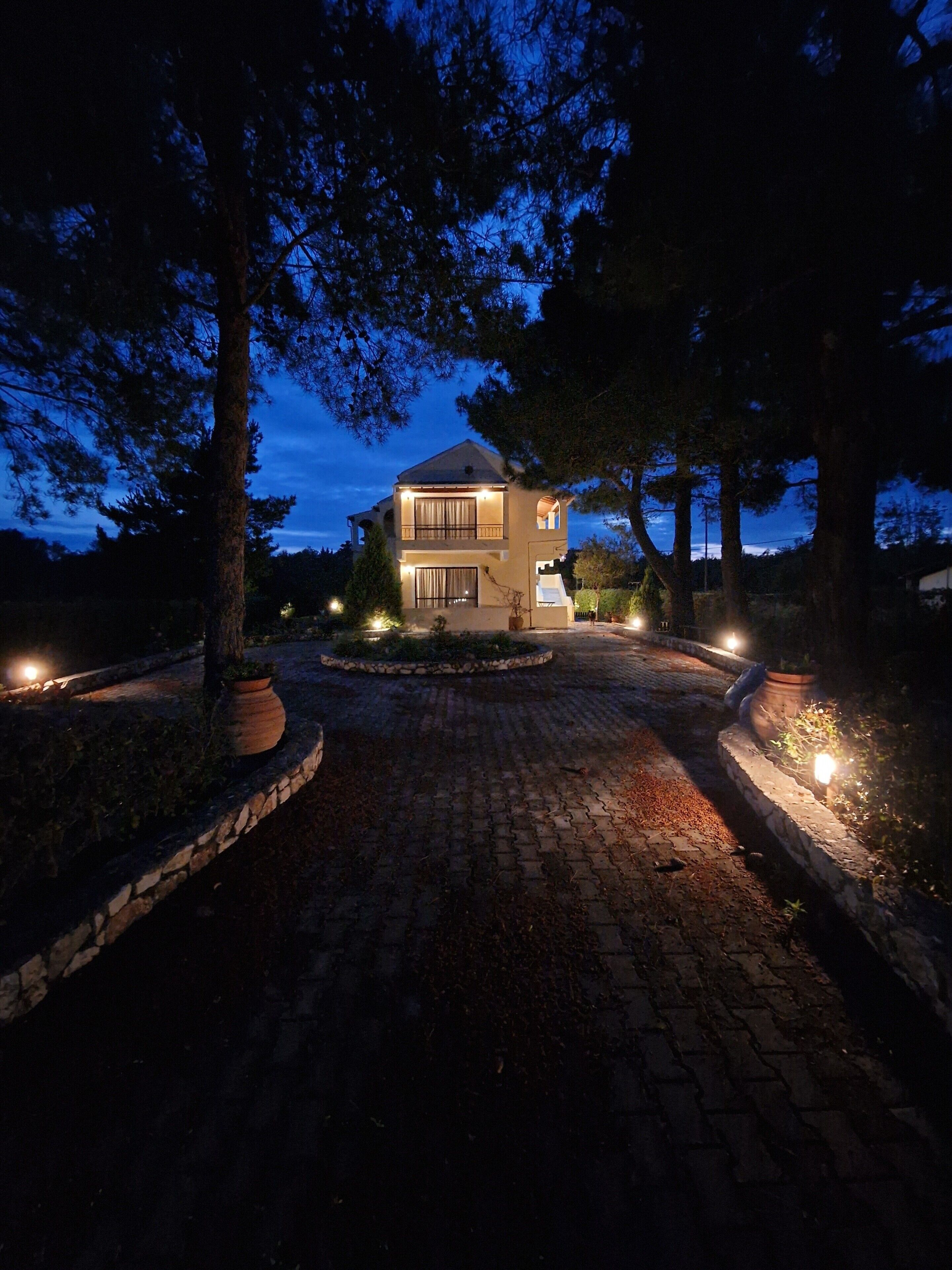A two-story house with a paved driveway, surrounded by trees, and illuminated by exterior lights.