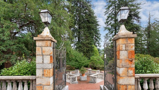 Une porte en pierre, agrémentée de barreaux métalliques, qui mène à une zone pavée avec un parterre central.
