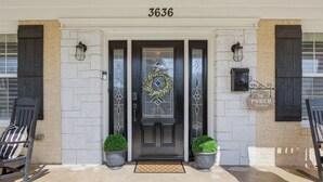 Una entrada principal con el número 3636, una puerta oscura adornada con un adorno navideño, dos plantas en macetas y un letrero que dice "The Porch Sit & Relax".