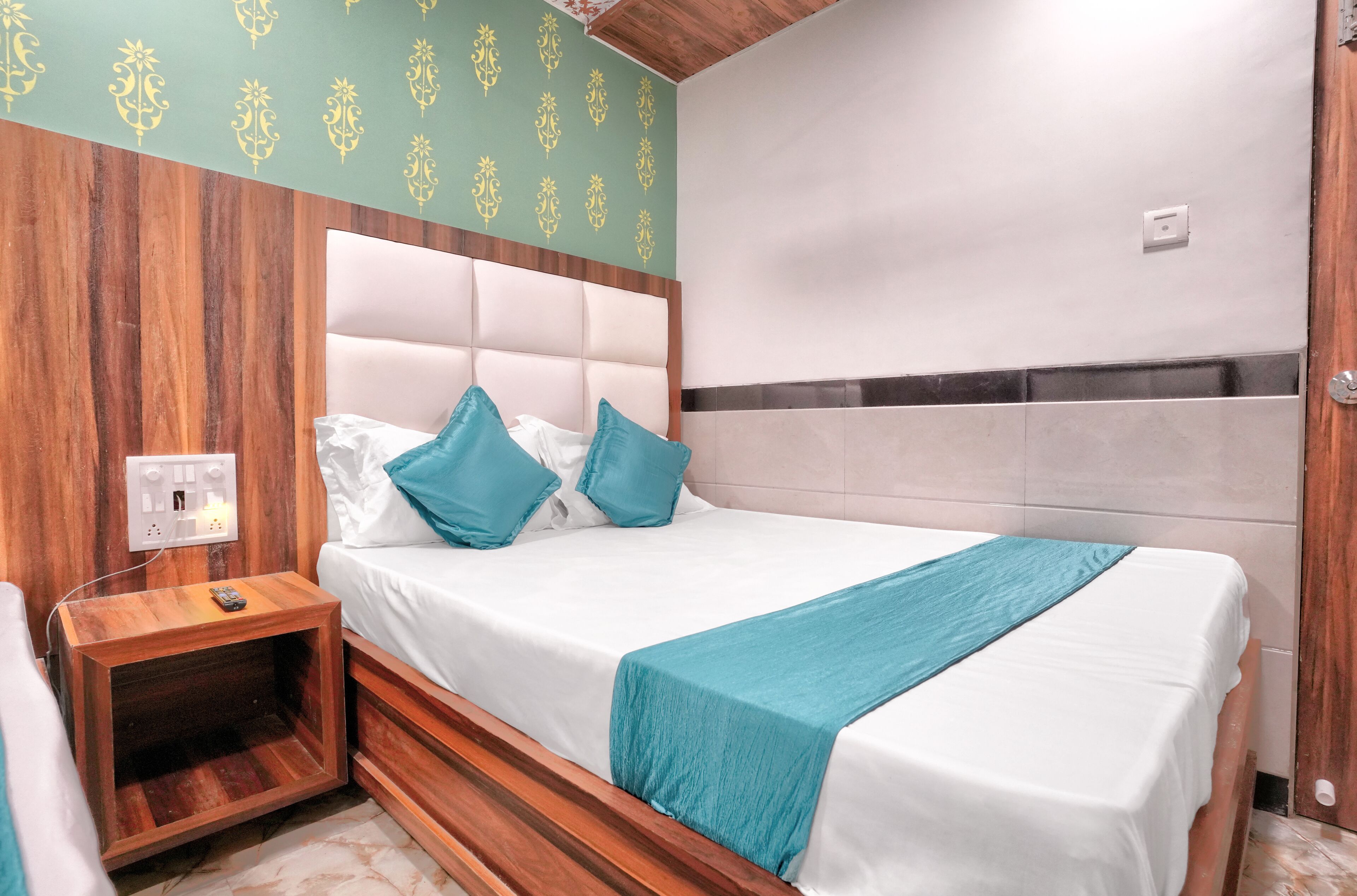 A neatly made bed with white and teal bedding, a wooden nightstand with a remote control, and a green patterned wall.