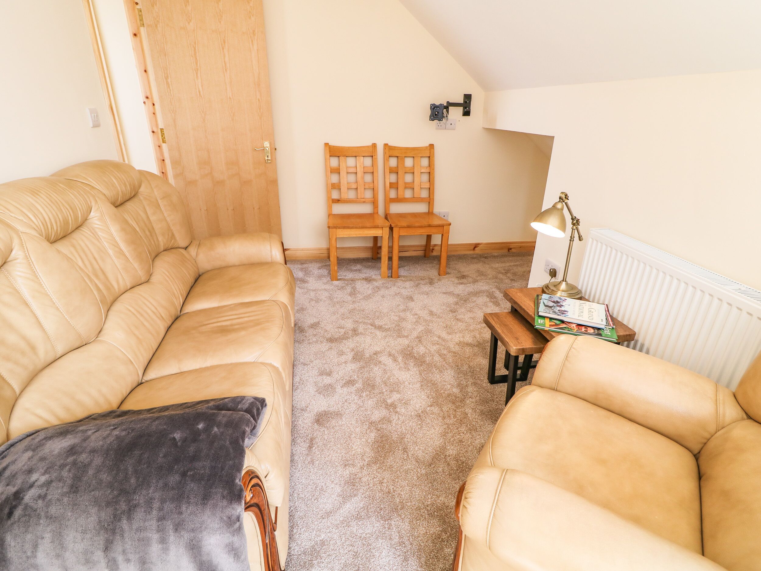 A cozy living room with a beige leather sofa, a dark throw pillow, and a wooden chair. A small wooden table holds a lamp and books. The room features a sloped ceiling and a radiator.