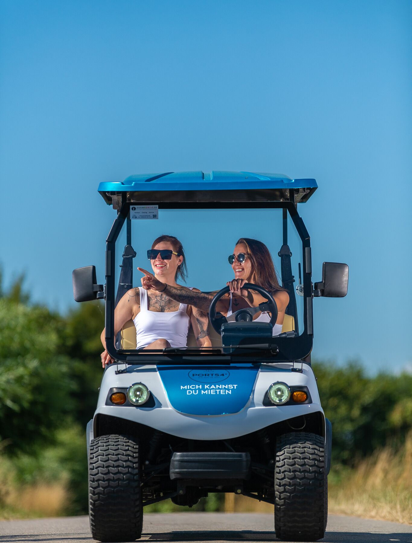 A golf cart with two people inside, driving on a road with greenery in the background.