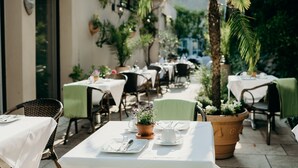 An outdoor dining area with tables set for two, surrounded by potted plants and chairs. - Hotel Crystal (Saarbrücken)