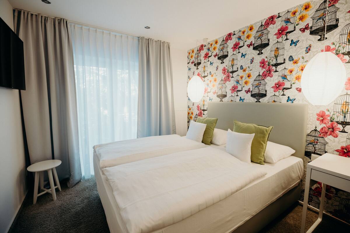 A hotel room with a large bed, a small stool, a TV, and a wall with a bird and flower pattern.