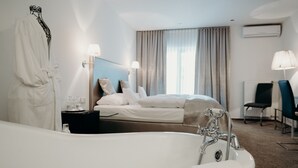 A hotel room with a large white bathtub, a bed with white linens, a bedside table with toiletries, a chair, and a window with curtains. - Hotel Crystal (Saarbrücken)