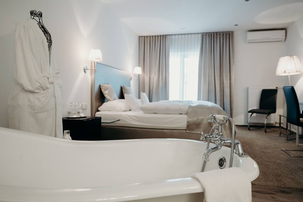 A hotel room with a large white bathtub, a bed with white linens, a bedside table with toiletries, a chair, and a window with curtains.