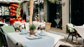 An outdoor dining area with tables set for two, featuring white tablecloths and a potted plant.