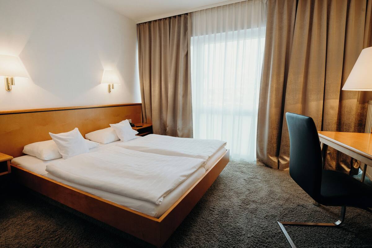 A hotel room with a double bed, a desk with a chair, a lamp, and a window with curtains.