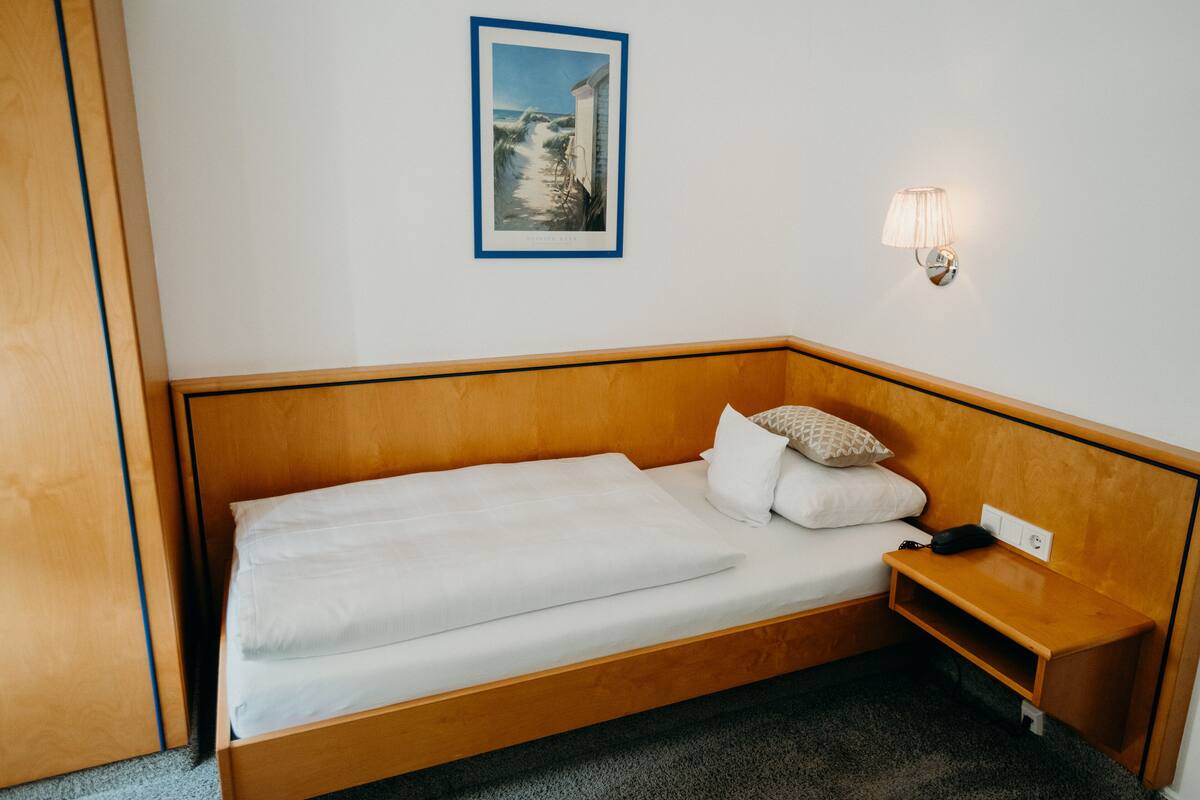 A single bed with a wooden headboard, a bedside table, a wall-mounted lamp, and a framed picture above the bed.