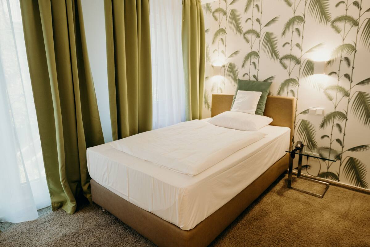 A neatly made bed with white linens in a room with green curtains and a wall with palm tree wallpaper.