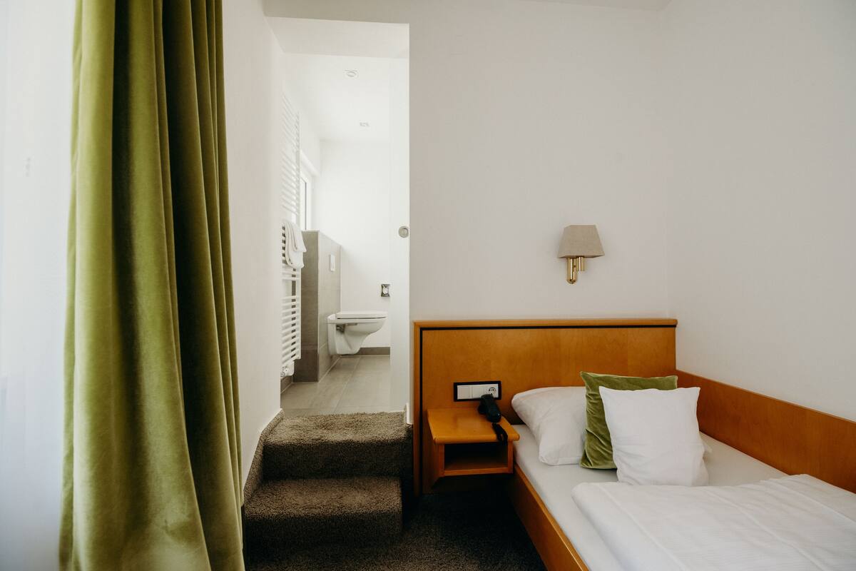 A hotel room with a bed, a nightstand, a lamp, a green curtain, and a view of a bathroom with a toilet and a shower.
