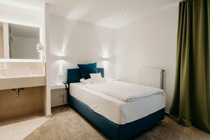 Una habitación de hotel moderna con una cama, una mesita de noche, un espejo y un baño con lavamanos y toallero.