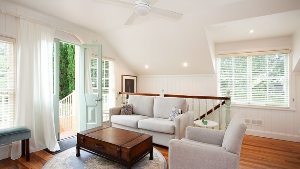 A spacious living room with a high ceiling, a ceiling fan, a light-colored sofa, a wooden coffee table, and a view of greenery outside.