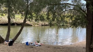 A sandy beach with a calm river, trees, and some camping gear.