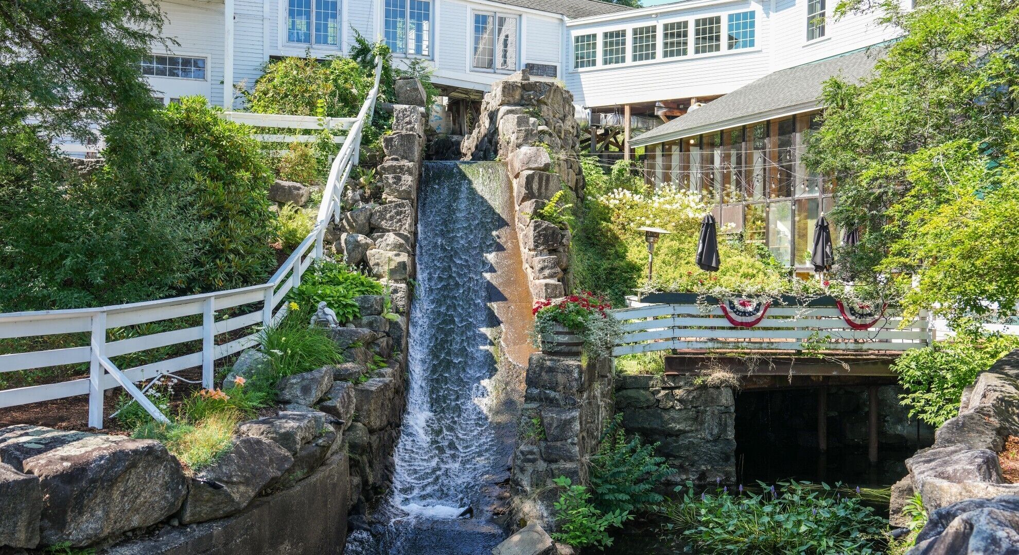 Une cascade ornée d’une clôture blanche, entourée d’une végétation luxuriante et d’un bâtiment doté de grandes fenêtres.