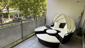 Ein Balkon mit einem Rattan-Sofa, einer überdachten Sitzgelegenheit, einem Metallgeländer und Blick auf einen Parkplatz.