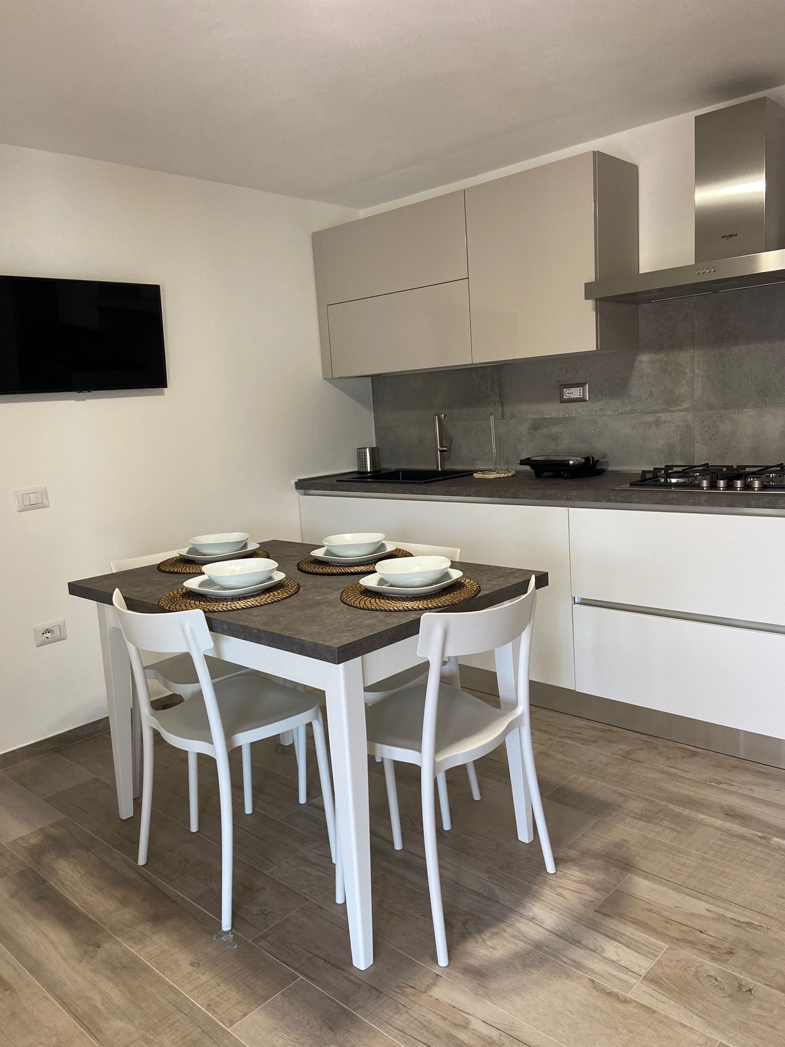 Une cuisine moderne avec des meubles blancs, un téléviseur à écran plat fixé au mur, une cuisinière à gaz et une table à manger avec des chaises blanches.