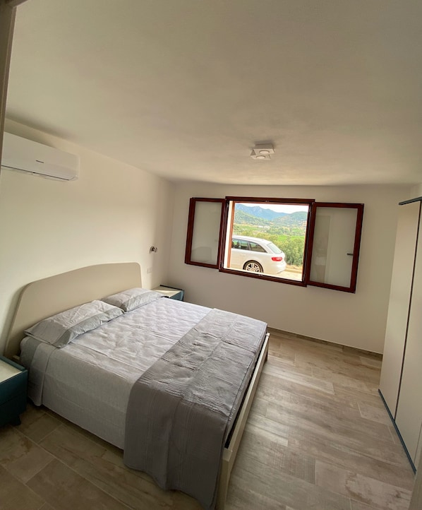 A bedroom with a bed, a nightstand, a window with a view of a car and landscape, and a wardrobe.