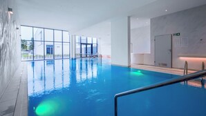 Piscina interna com paredes de vidro transparente, sinalização de saída visível e símbolo de sauna na parede.