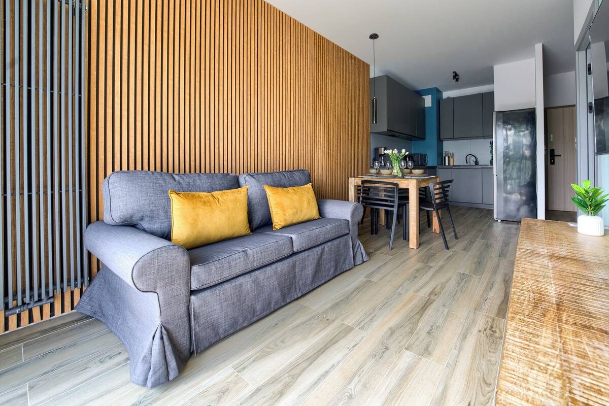 A modern living room with a grey sofa, wooden slatted wall, and a dining area with a wooden table and chairs.