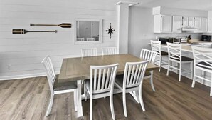 A dining area with a wooden table and white chairs, a white wall with a compass decoration, and a kitchen area with white cabinets.