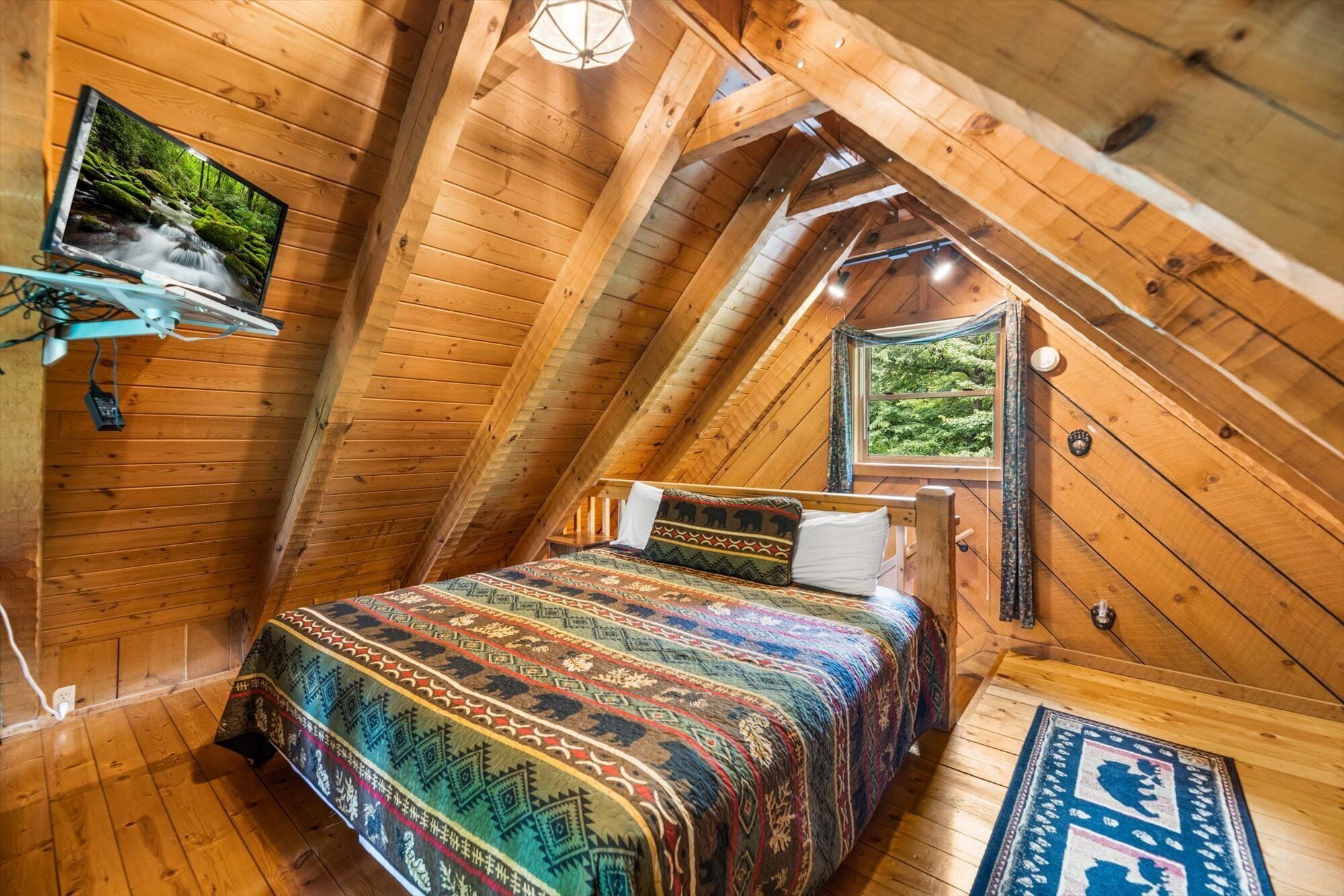 A cozy attic bedroom with a wooden sloped ceiling, a bed with patterned bedding, a mounted television, and a window with a view of greenery.
