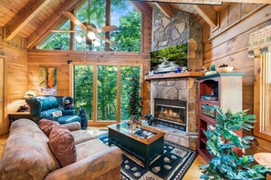 Interior de una cabaña de troncos con chimenea, muebles de madera y ventanas amplias que dan a un bosque.