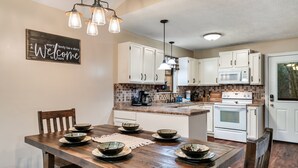 A kitchen with white cabinets, a wooden dining table with bowls, and a 'Welcome' sign. - Deer Browse Hideaway: Sink a Putt, Soak It In (Sevierville)