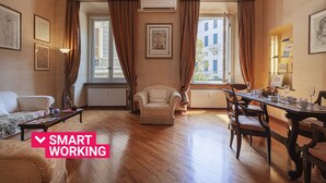 Ein geräumiges Zimmer mit Essbereich, einer Couch und einem Sessel. Vor den Fenstern hängen Vorhänge und die Wände sind mit Kunstwerken geschmückt.