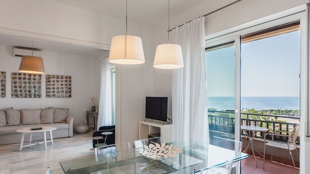 A modern living room with a glass dining table, a sofa, and a TV on a stand. There is a balcony with a view of the sea. - Piazza Europa Landscape by Wonderful Italy (Catania)