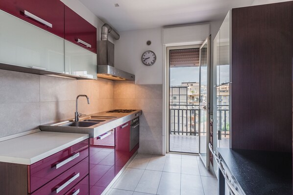 Une cuisine compacte équipée d’une cuisinière à gaz, d’un évier et de meubles de rangement modernes. Une horloge est visible au mur, et la cuisine donne sur un balcon avec vue sur la ville.