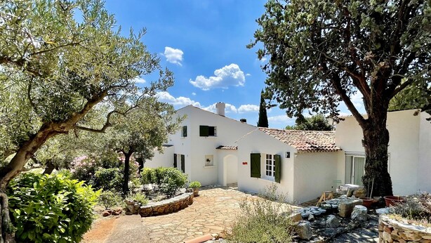 Un bâtiment blanc avec un toit en tuiles, entouré de verdure et bordé d’un chemin en pierre.