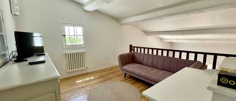 A room with a television on a stand, a radiator, a brown sofa, a round rug, and a white desk.