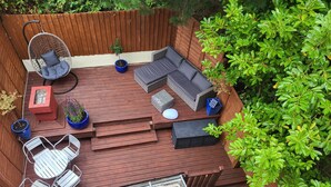 A wooden deck with a built-in seating area, including a sofa and chairs, surrounded by greenery. - Beach Front 2-bed Apartment With Deck & BBQ (Bournemouth)