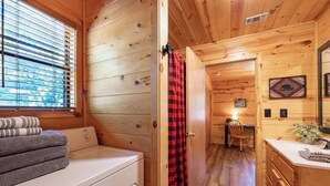 L’intérieur d’une cabane en bois comprend une machine à laver, un rideau à carreaux rouges et noirs, une table à manger en bois avec une chaise, et un panneau indiquant les règles concernant le séjour dans la cabane.