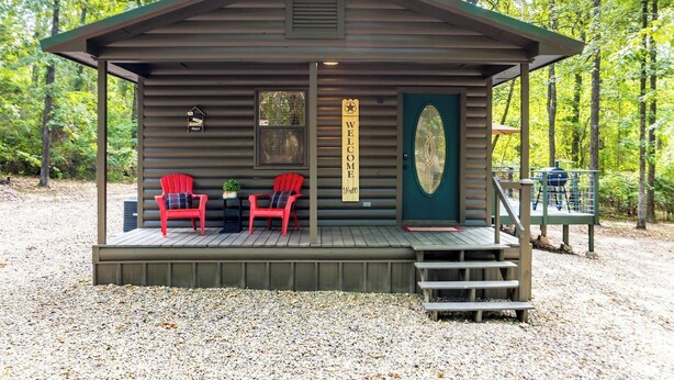 Une cabane avec une véranda, deux chaises Adirondack rouges, une pancarte de bienvenue et un barbecue.