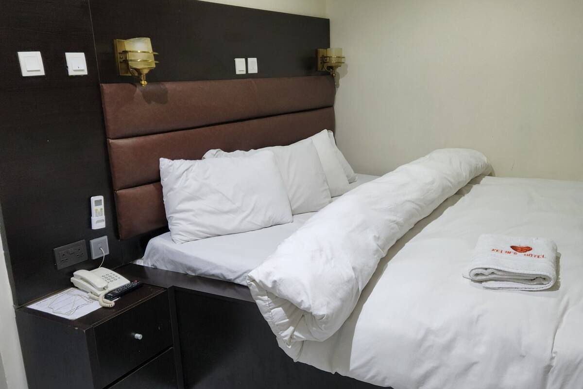 A neatly made bed with white linens, a bedside table with a phone and a lamp, and a painting on the wall.