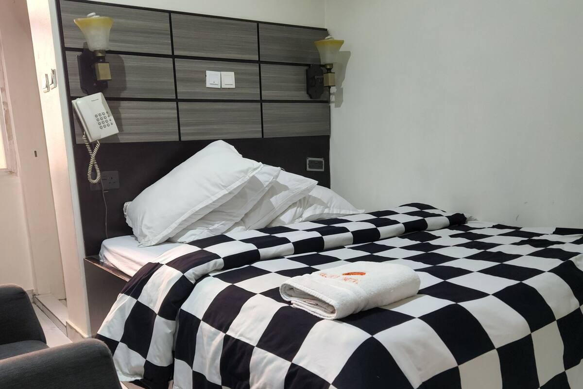 A hotel room with a checkered bedspread, a bedside lamp, a telephone, and a chair.