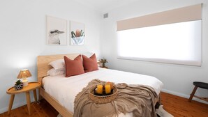 A bedroom with a wooden bed, white bedding, a bedside table with a lamp, and a window with a blind. - Quay Haven – complete relaxation & fun (Callala Beach)