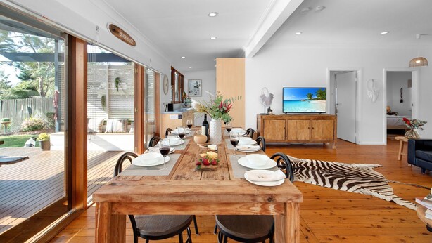 A dining area with a wooden table set for four, a television, and a view of the outdoors. - Quay Haven – complete relaxation & fun (Callala Beach)