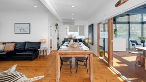 A modern living space with a dining area, a black leather sofa, and a wooden dining table set for six. - Quay Haven – complete relaxation & fun (Callala Beach)