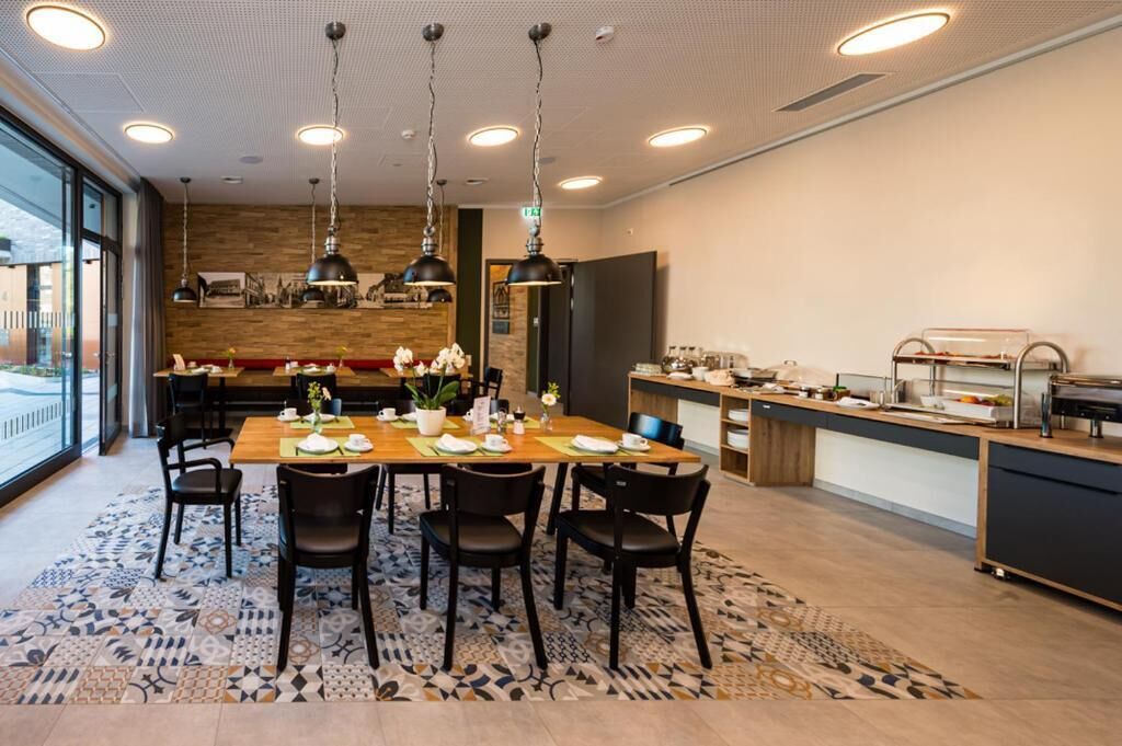 Un comedor moderno con una mesa de madera dispuesta para cuatro personas, rodeada de sillas negras. Hay lámparas colgantes sobre la mesa y una alfombra estampada en el suelo.