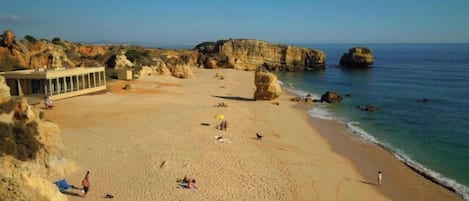 Uma praia com céu limpo, falésias rochosas e algumas pessoas.
