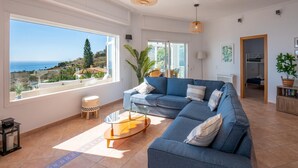 Ein geräumiges Wohnzimmer mit großem Fenster und Blick auf das Meer und die Hügel.